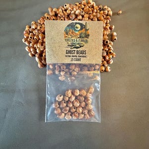 May include: A clear plastic bag containing small, round, light brown natural juniper and cedar beads, labeled Ghost Beads with a Cactus & Timber Trading Company logo and 25 count text, surrounded by a loose pile of identical brown wooden beads.