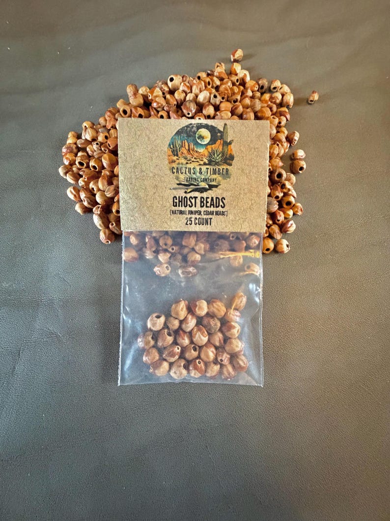 May include: A clear plastic bag containing small, round, light brown natural juniper and cedar beads, labeled Ghost Beads with a Cactus & Timber Trading Company logo and 25 count text, surrounded by a loose pile of identical brown wooden beads.