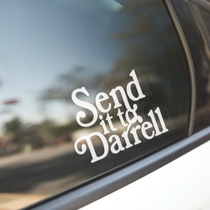 May include: White vinyl decal with the phrase "Send it to Darrell" in a decorative font, affixed to a car window. The background shows a blurred street scene with trees and buildings.