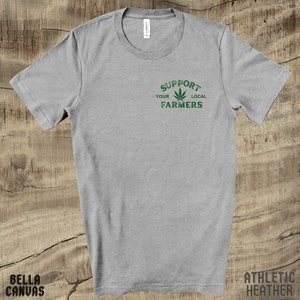 K&ouml;nnte beinhalten: Ein graues T-Shirt mit dem Text "Support Your Local Farmers" in gr&uuml;ner Schrift mit einer gr&uuml;nen Marihuana-Blattgrafik.