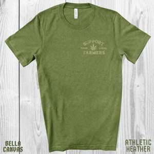 K&ouml;nnte beinhalten: Gr&uuml;nes T-Shirt mit einer Grafik eines Marihuana-Blattes und dem Text "Unterst&uuml;tze deine lokalen Bauern".