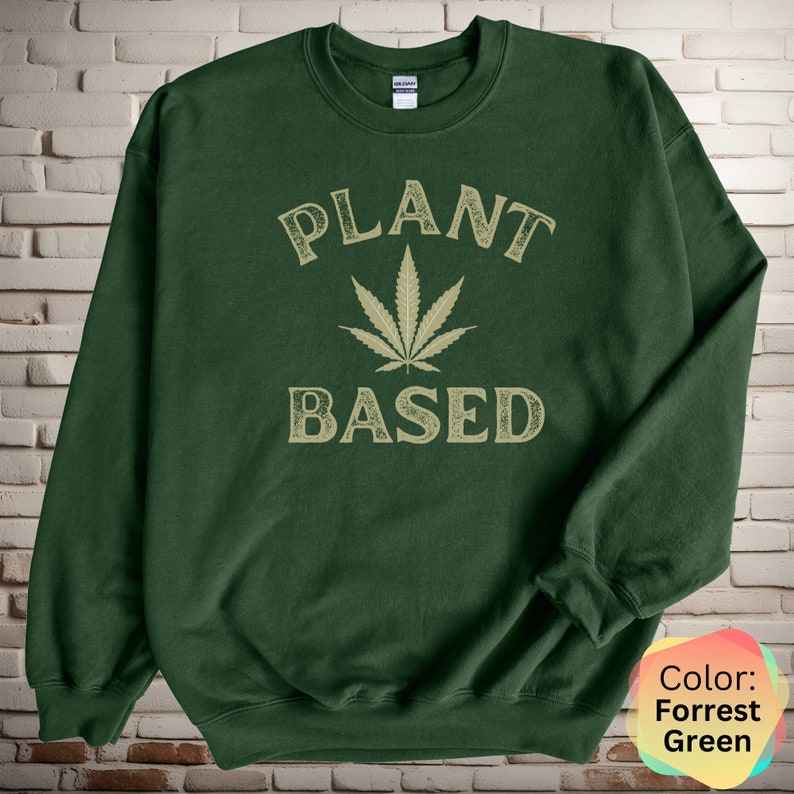 Puede incluir: Una sudadera verde oscuro con el texto "PLANT BASED" en una fuente de estilo vintage. Una hoja de marihuana estilizada se encuentra entre las palabras.