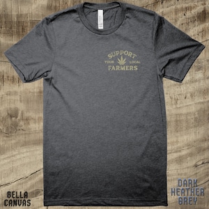 K&ouml;nnte beinhalten: Dunkelgraues T-Shirt mit einem goldenen Aufdruck, der "Unterst&uuml;tze deine lokalen Landwirte" mit einem Marihuana-Blatt-Symbol sagt.