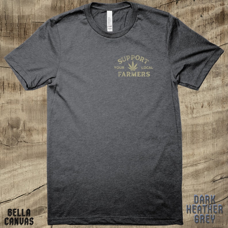 K&ouml;nnte beinhalten: Dunkelgraues T-Shirt mit einem goldenen Aufdruck, der "Unterst&uuml;tze deine lokalen Landwirte" mit einem Marihuana-Blatt-Symbol sagt.