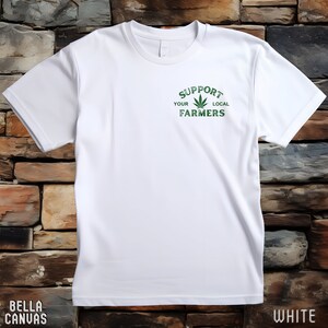 K&ouml;nnte beinhalten: Wei&szlig;es T-Shirt mit einem gr&uuml;nen Marihuana-Blatt-Motiv und dem Text "Unterst&uuml;tze deine lokalen Bauern".