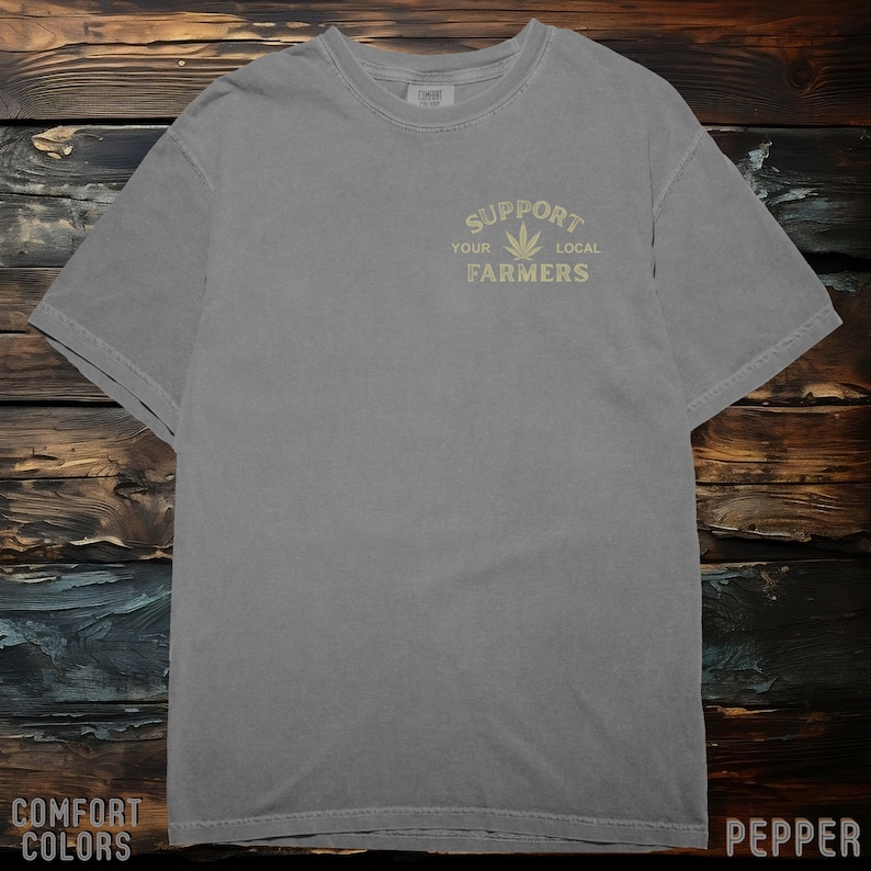 K&ouml;nnte beinhalten: Ein graues T-Shirt mit einer Grafik im Vintage-Stil, auf dem "Unterst&uuml;tze deine lokalen Bauern" mit einem Marihuana-Blatt-Symbol steht.