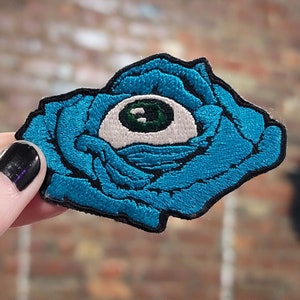 May include: A blue embroidered patch of a rose with a white eye in the centre. The eye has a green iris and a black pupil.