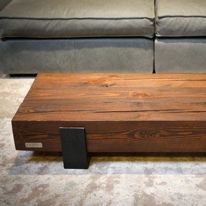 Op de afbeelding: Een rechthoekige, donkerbruine houten salontafel met zichtbare houtnerf. De tafel wordt ondersteund door twee zwarte metalen beugels. De tafel staat voor een grijze bank.