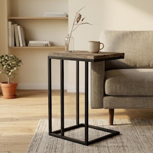 Op de afbeelding: Een C-vormige bijzettafel met een rechthoekig houten blad met een verweerde bruine afwerking en een zwart metalen frame. De tafel staat naast een beige bank en bevat een kleine doorzichtige glazen vaas met een plant en een keramische mok.