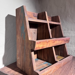 May include: A tiered wooden display rack featuring a natural brown finish with visible wood grain and distressed accents in shades of blue, green, and yellow. The structure includes two levels with open compartments, angled side panels, and a sturdy base.