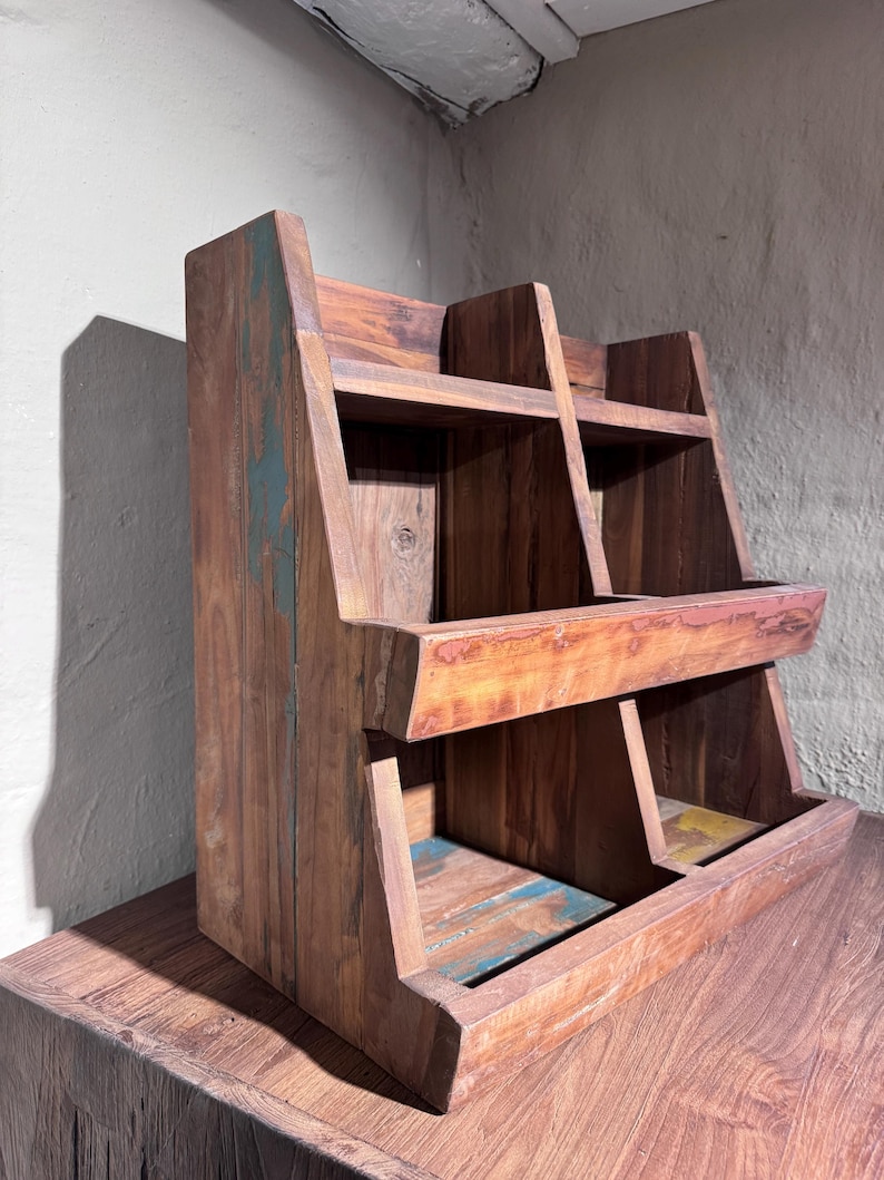 May include: A tiered wooden display rack featuring a natural brown finish with visible wood grain and distressed accents in shades of blue, green, and yellow. The structure includes two levels with open compartments, angled side panels, and a sturdy base.