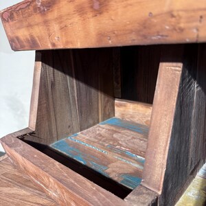 May include: A tiered wooden display stand featuring a dark brown finish with visible wood grain. The structure includes an open shelf with a base showing patches of weathered blue paint, and a top section with a worn, reddish-brown stained edge.