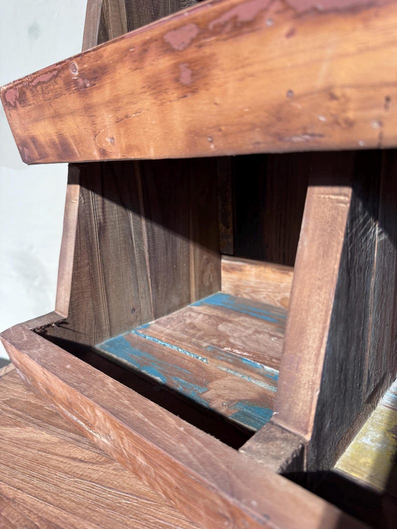 May include: A tiered wooden display stand featuring a dark brown finish with visible wood grain. The structure includes an open shelf with a base showing patches of weathered blue paint, and a top section with a worn, reddish-brown stained edge.
