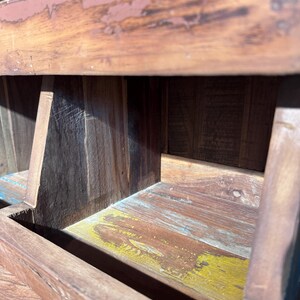 May include: A small wooden step stool featuring a weathered brown finish with visible wood grain. The structure includes a flat top surface and a lower step, with hints of yellow and blue paint showing through the distressed wood texture.