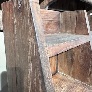May include: A tiered wooden shelf unit featuring a dark brown finish with visible wood grain patterns. The structure consists of two open shelves with angled side panels, showcasing a rustic texture and a sturdy, multi-level design for storage.