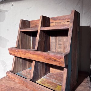 May include: A tiered wooden display shelf featuring two levels with two open compartments on each level. The structure is crafted from reclaimed wood with visible grain, brown tones, and subtle hints of blue and yellow paint on the base.