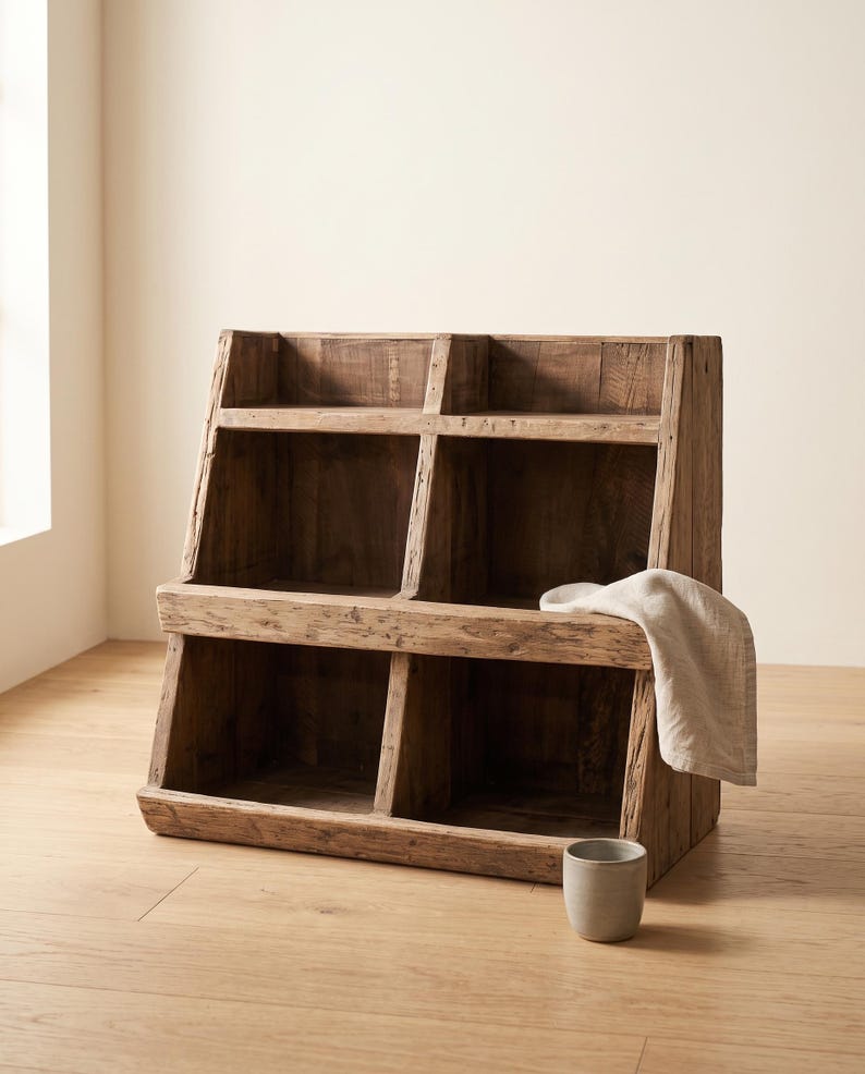 May include: A rustic wooden storage unit featuring four open compartments arranged in two tiers. The structure is crafted from weathered brown wood with visible grain. A light-colored fabric cloth is draped over the side, and a small gray cup sits.
