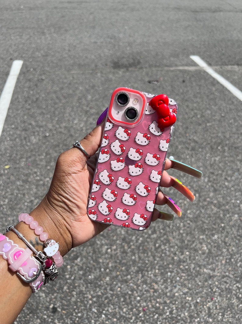 May include: A clear phone case with a pink background and a repeating pattern of Hello Kitty faces. The case has a red camera ring and a red Hello Kitty bow charm.