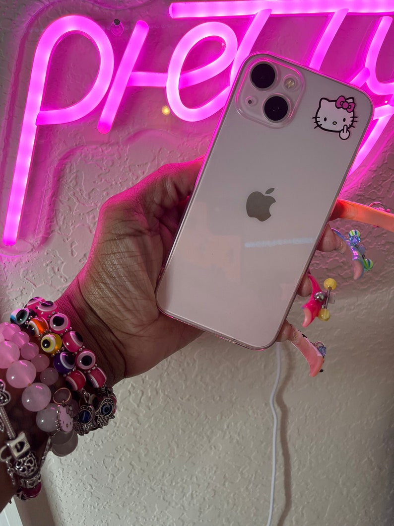 May include: A white iPhone with a Hello Kitty sticker on the back. The phone is being held by a hand with several bracelets on the wrist.