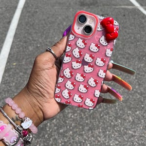 May include: A pink clear phone case with a Hello Kitty design and a red bow charm. The case has a clear back and a pink Hello Kitty pattern. The case has a red bow charm on the back.