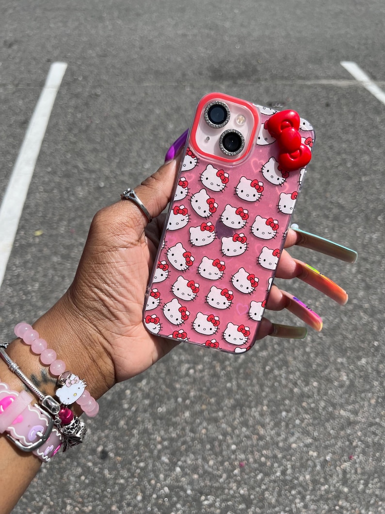 May include: A pink clear phone case with a Hello Kitty design and a red bow charm. The case has a clear back and a pink Hello Kitty pattern. The case has a red bow charm on the back.