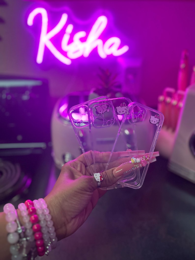 May include: Three clear phone cases with a Hello Kitty design. The cases are stacked on top of each other and are being held in a hand.