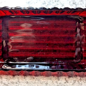 May include: A rectangular ruby red glass butter dish with a ribbed interior base and scalloped edges. The dish features small, rounded handles on each end with a sunburst pattern, and the overall design includes a decorative scalloped rim.