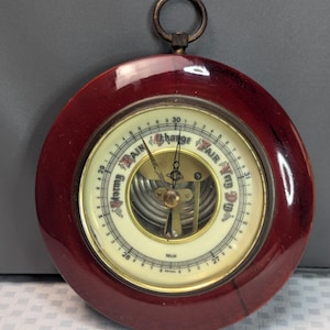 May include: A round analog barometer with a polished dark reddish-brown wooden frame and a brass hanging ring. The cream-colored dial features black text reading Stormy, Rain, Change, Fair, and Very Dry, with numerical scales and two metal hands.