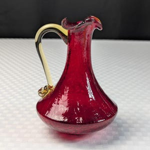 May include: A small, ruby red glass pitcher with a curved handle. The handle is cream-colored with black stripes and gold accents. The pitcher has a flared spout and a bulbous base, perfect for serving beverages.