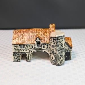 May include: A ceramic miniature building figurine featuring a textured stone-look facade in shades of gray and white, topped with a glossy orange-brown tiled roof. The structure includes a prominent cylindrical tower and arched openings.