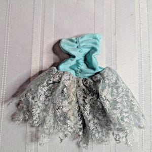 May include: A miniature doll dress featuring a light blue bodice and a silver lace skirt. The bodice is gathered at the neckline. The skirt is made of delicate, layered lace. The dress is designed for a doll.
