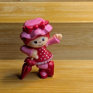 May include: A small plastic figurine of a character wearing a pink hat with a bow, a red dress with white polka dots, and pink boots. The character has red hair, a smiling face, and is holding a closed dark red umbrella in one hand.