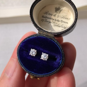 May include: Diamond stud earrings presented in an open, antique black jewellery box with a blue velvet lining. The box features gold detailing and the inscription "Brock & Sons, Goldsmiths to the Queen, 87 George St. Edinburgh."