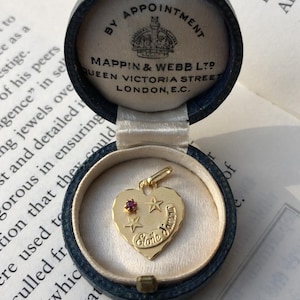May include: A gold heart pendant with a ruby and star details, displayed in an open blue velvet box. The pendant is inscribed with "Etoile Jamais". The box is from Mappin & Webb, Queen Victoria Street, London.