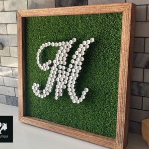 May include: Square wooden framed wall art featuring a green artificial grass background. The letter "M" is crafted from numerous small white objects. The frame is a light brown color. The Back9Boutique logo is in the lower left corner.