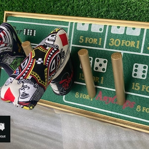 May include: A pair of high-heeled shoes with playing card designs, displayed on a green felt surface with gold trim. The felt has printed dice and text. The shoes have a colorful, patterned design.