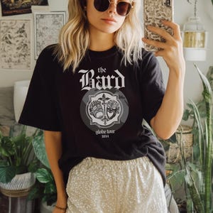 May include: Black t-shirt with the text "the Bard" in a gothic font, and a graphic of theater masks. The shirt also has the text "globe tour 1614". The person is wearing gold-rimmed sunglasses and sequin shorts.
