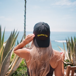 May include: A black baseball cap with the words "Thinking Cap" embroidered in yellow on the front. The cap is worn by a person with long blonde hair, who is looking out at the sea.