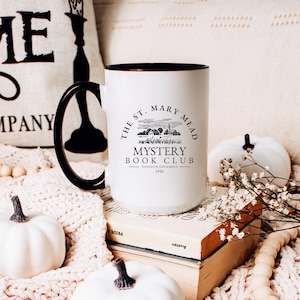 May include: White ceramic coffee mug with a black handle and black rim. The mug has a black and white graphic of a village with the text "The St. Mary Mead Mystery Book Club Founded by Jane Marple 1930."