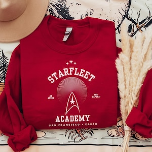 Puede incluir: Una sudadera roja de Starfleet Academy con texto y gráficos blancos. El diseño incluye las palabras "Starfleet Academy" y la insignia de Starfleet. También se incluye el texto "Ad Astra" y "Per Aspera". La sudadera está doblada sobre una superficie estampada.