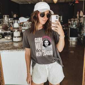 Peut inclure: T-shirt gris anthracite avec l'inscription "GIRL MATH LIKE HEDY LAMARR" et un portrait d'Hedy Lamarr. Le t-shirt est associé à un short à carreaux blancs et gris et à une casquette de baseball blanche.
