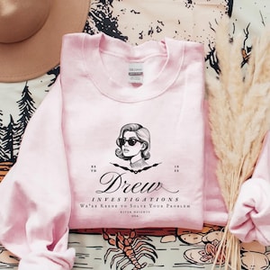 May include: Pink sweatshirt with a black and white illustration of a woman wearing sunglasses and the text "Drew Investigations We're Keene to Solve Your Problem River Heights USA Est. 1935"