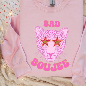 May include: A light pink sweatshirt with the words "BAD BOUJEE" in pink and gold lettering. The design features a pink leopard face wearing gold star-shaped sunglasses. The sweatshirt is a crew neck style.