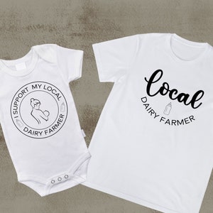 May include: White baby onesie with the text "I support my local dairy farmer" in a circle with a breastfeeding mother and a milk bottle graphic. White t-shirt with the text "local dairy farmer" in cursive with a milk bottle graphic.