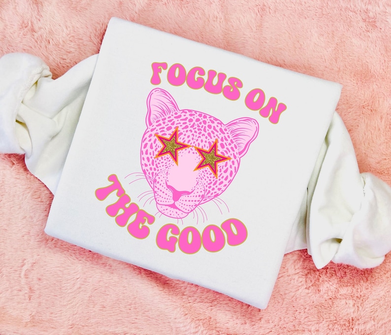 May include: White t-shirt with a pink leopard print design wearing gold star sunglasses. The text "FOCUS ON THE GOOD" is written in pink with a gold outline.