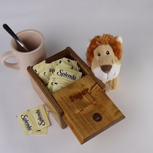 May include: A wooden box with the word Sugar engraved on the lid, filled with yellow Splenda packets. A light pink mug with a black spoon, two loose Splenda packets, and a small plush lion toy are positioned around the wooden container.