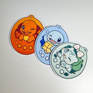 Könnte beinhalten: Drei farbenfrohe Aufkleber mit Zeichentrickfiguren. Die Aufkleber zeigen ein Charmander in einem orangefarbenen Oval, ein Schiggy in einem blauen Oval und ein Bisasam in einem grünen Oval. Jede Figur lächelt und ist von einem dekorativen Rand umgeben.