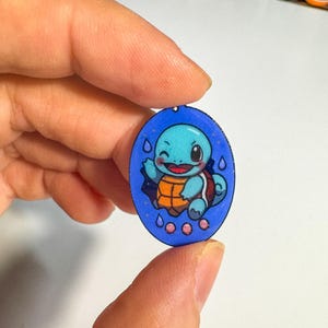Könnte beinhalten: Blauer, ovaler Anhänger mit einem Cartoon-Squirtle-Charakter mit lächelndem Gesichtsausdruck. Der Anhänger hat einen blauen Rand und ein kleines Loch zum Aufhängen. Squirtle ist blau, mit einem orangefarbenen Panzer und weißen Akzenten.