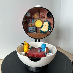 Könnte beinhalten: Ein 3D-gedruckter Pokéball mit einem Relaxo im Inneren, der auf einem Tisch steht. Der Pokéball ist geöffnet und zeigt Pikachu und Bisasam, die an einem Tisch ein Videospiel spielen, auf dem die Worte "GAME OVER" auf dem Bildschirm zu sehen sind.