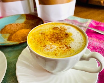 May include: A white ceramic mug filled with a golden yellow beverage topped with a layer of white foam and sprinkled with cinnamon. The mug is sitting on a white saucer.
